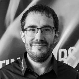 Homme souriant portant des lunettes et une barbe, devant un décor d'événement flou.