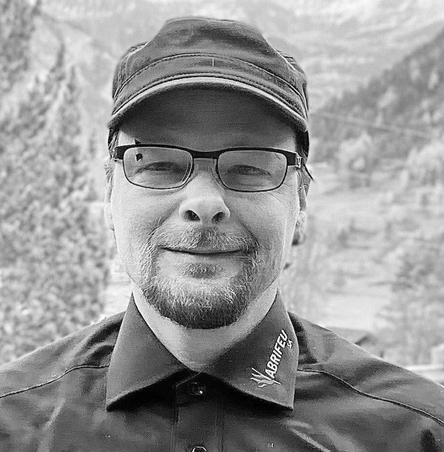 Un homme portant des lunettes et une casquette sourit en extérieur, devant un paysage de montagne.