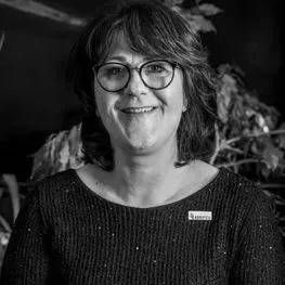Personne souriante portant des lunettes et un pull foncé sur fond intérieur flou