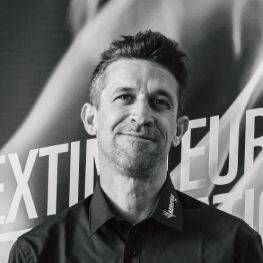 Un homme en chemise sombre sourit devant un fond recouvert de texte.