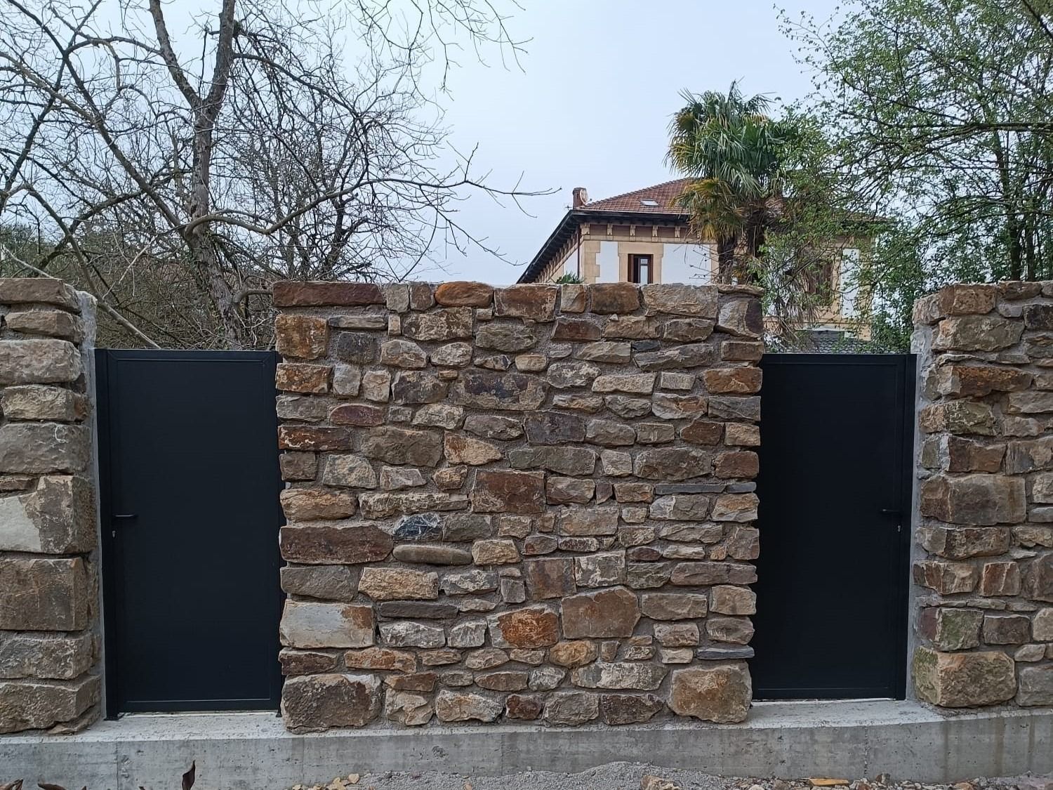 Un muro de piedra con una puerta negra y una casa al fondo.