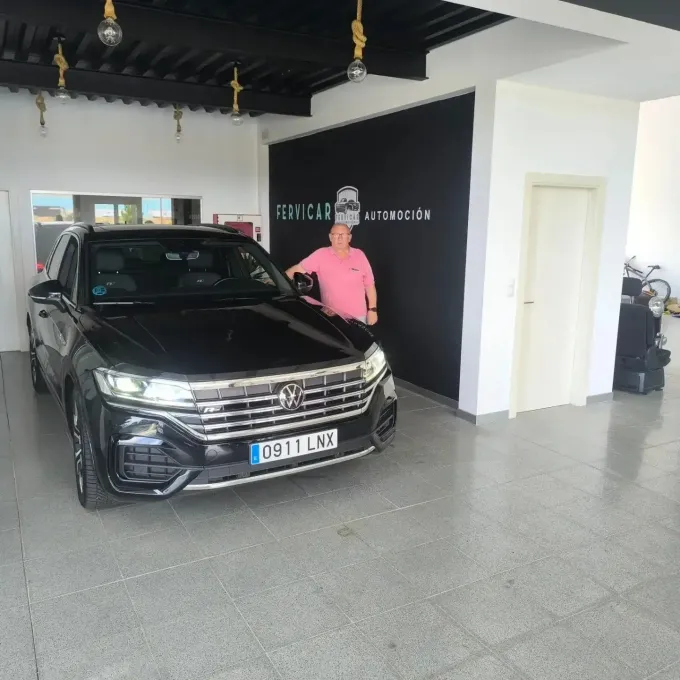 Un Volkswagen negro en una sala de exposición, con una persona vestida con una camisa rosa de pie junto al coche.