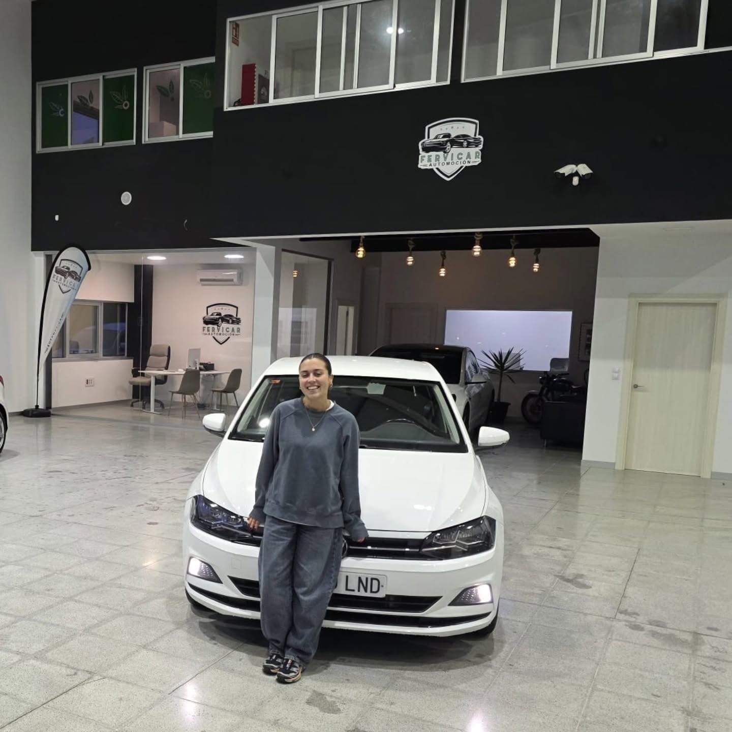 Persona de pie frente a un coche blanco dentro de una sala de exposición con suelo de baldosas y paredes negras.