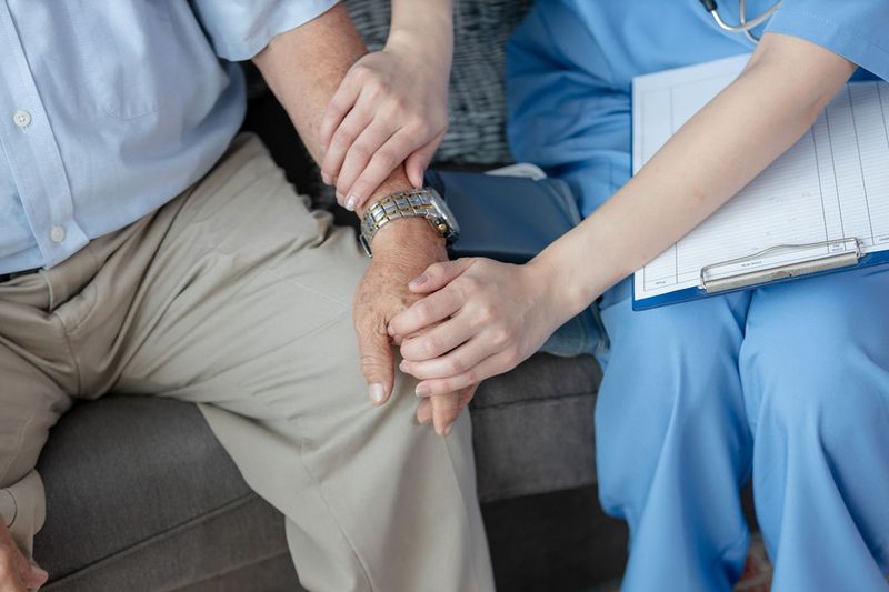 Une infirmière en blouse bleue réconforte un homme âgé en le tenant par le bras.