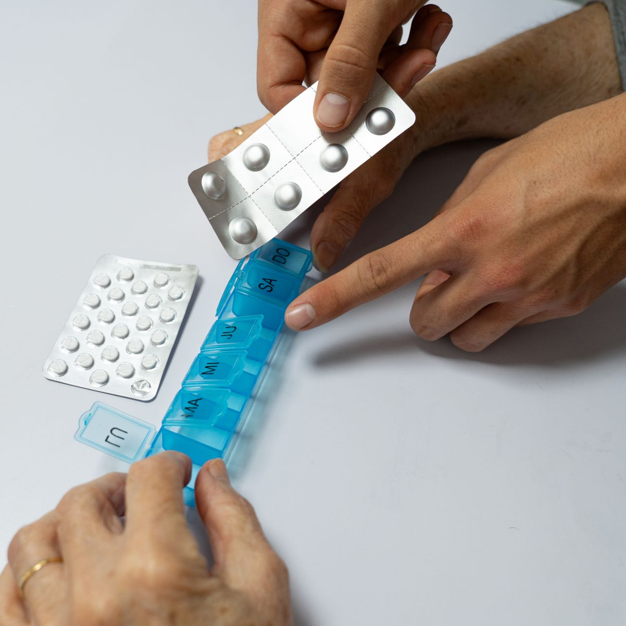 Des mains tiennent des plaquettes de médicaments et un pilulier bleu au-dessus d'une table blanche.