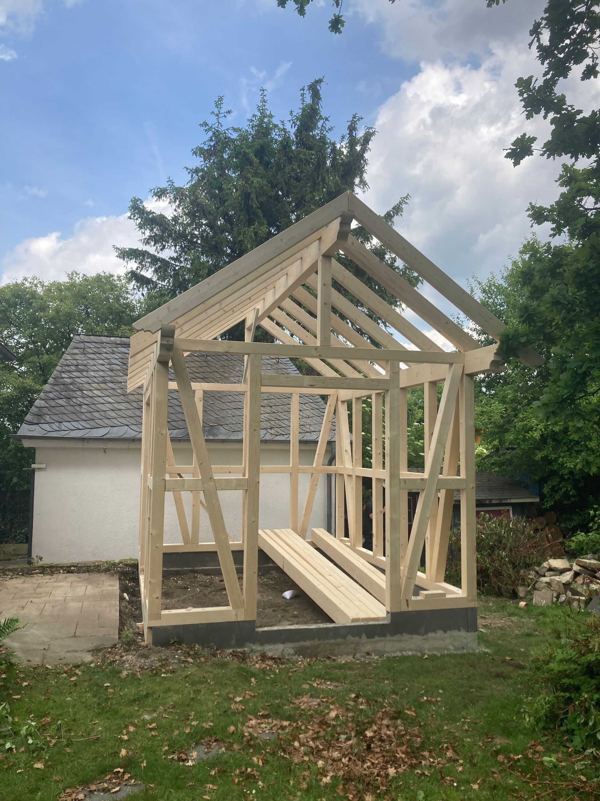 Holzrahmen eines kleinen, im Bau befindlichen Gebäudes, im Hintergrund ist ein teilweise sichtbares, bereits bestehendes Gebäude zu erkennen.
