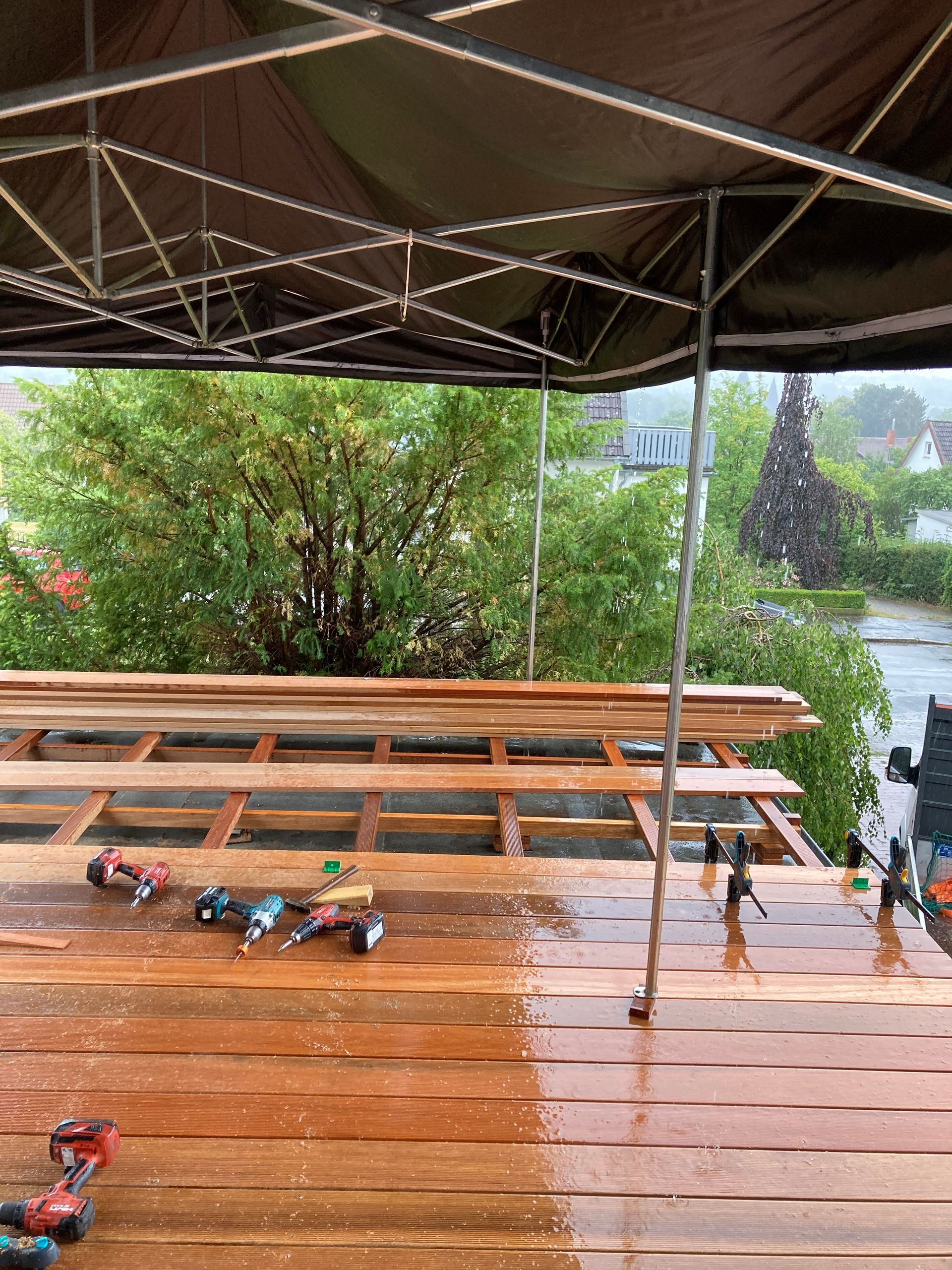 Eine verregnete Baustelle im Freien. Werkzeuge auf der Terrasse unter einem Zelt. Grüne Bäume und ein Haus im Hintergrund.