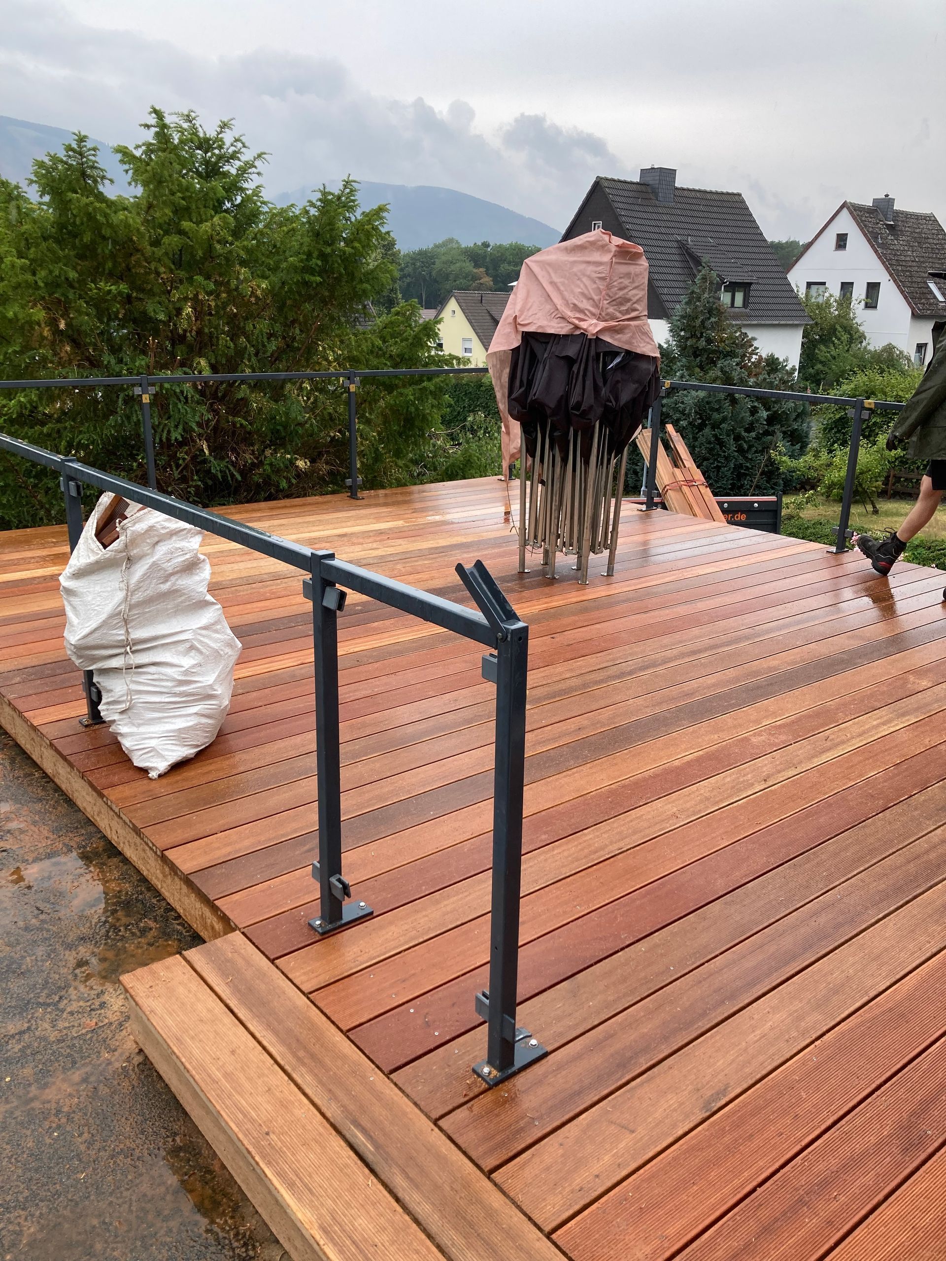 Holzterrasse mit schwarzem Geländer, Skulptur mit rosa Plane bedeckt, in der Ferne Gebäude und Berge.