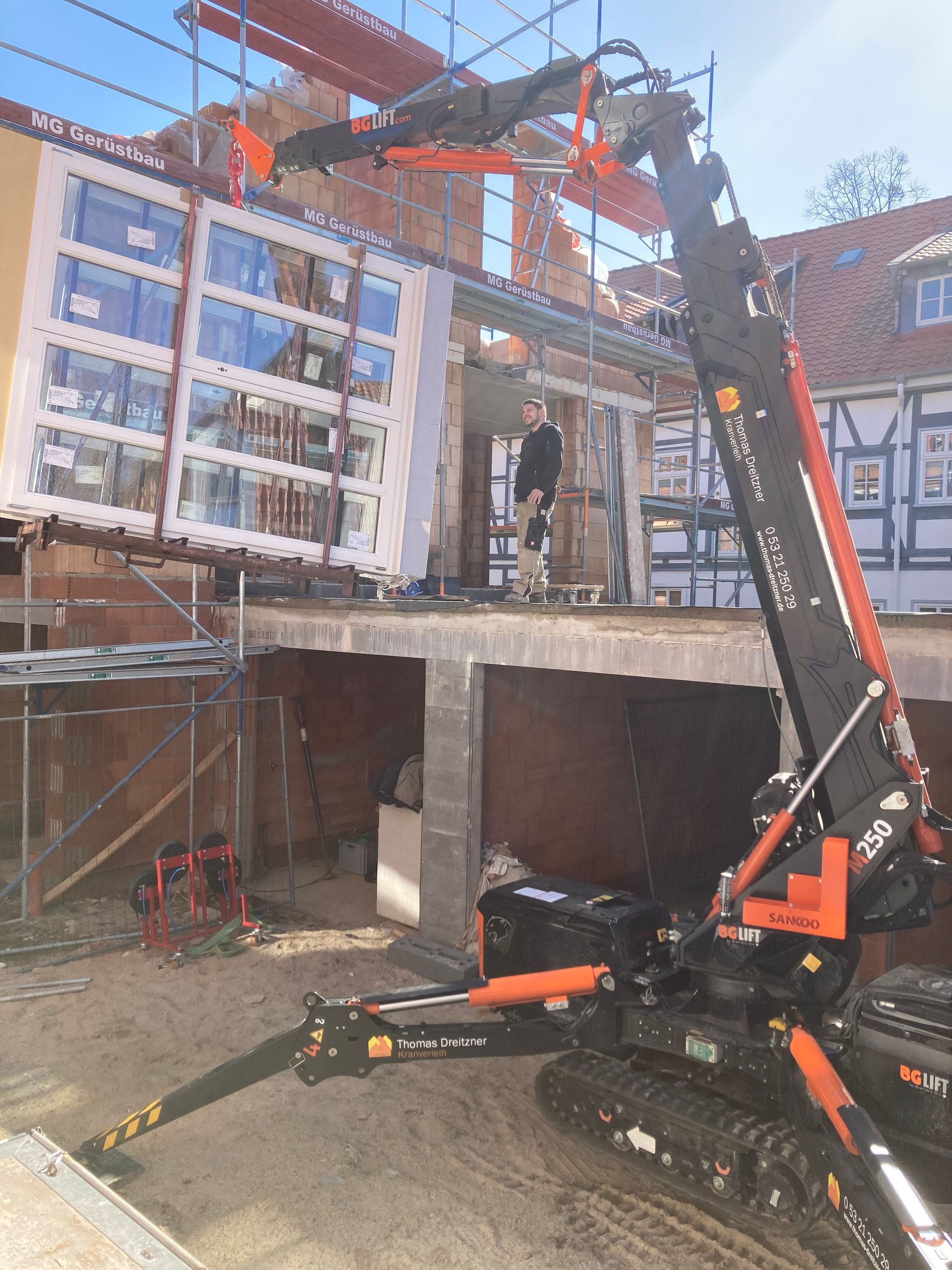 Ein Mobilkran hebt eine große Fenstereinheit in ein im Bau befindliches Gebäude. Ein Gerüst ist sichtbar.