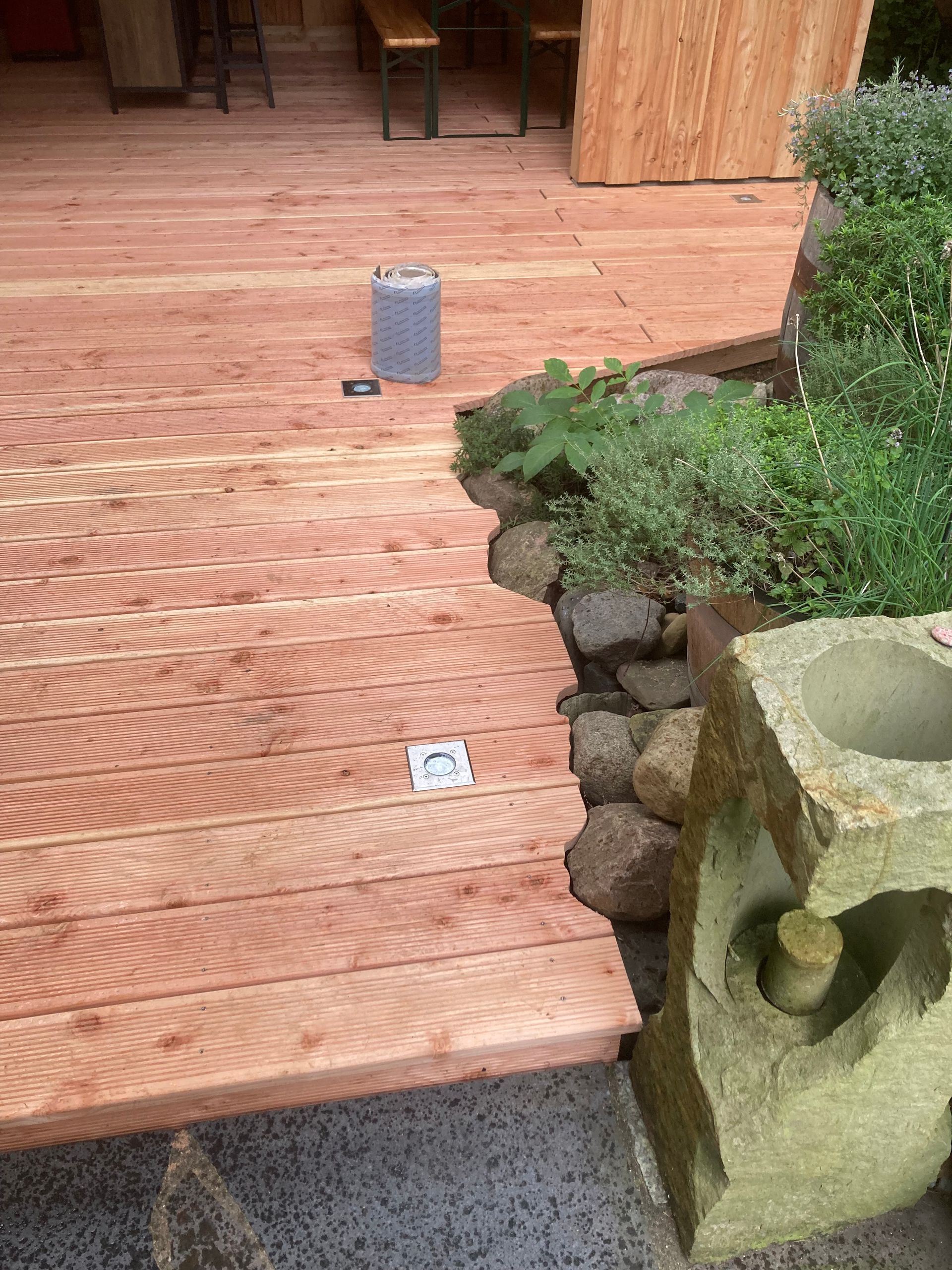 Holzterrasse mit unregelmäßiger Kante, angrenzend an eine Steinmauer mit Pflanzen und einer Steinskulptur.