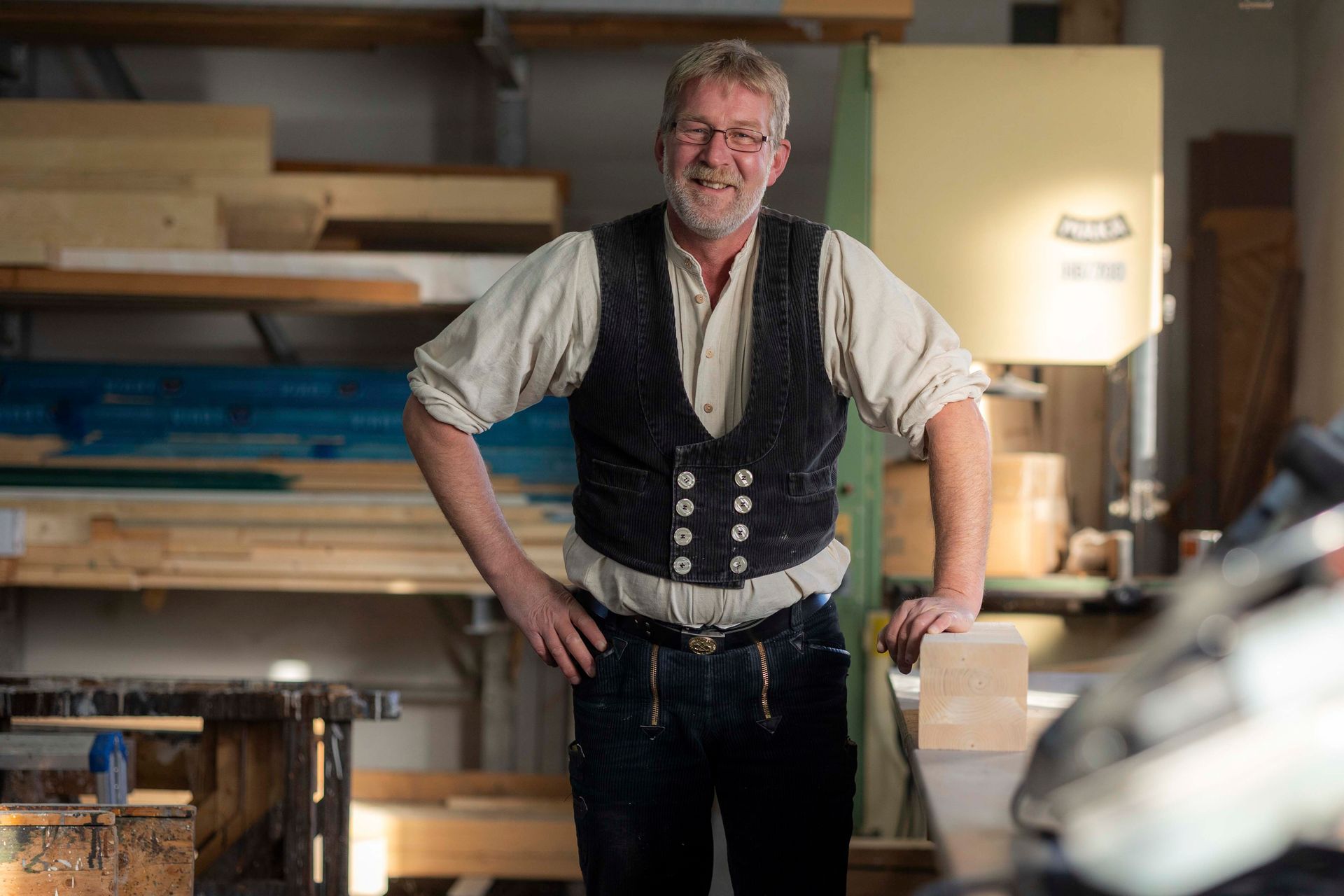 Ein Mann in einer Werkstatt lächelt, trägt eine Weste und lehnt an einem Holzblock. Im Hintergrund sind Regale und Maschinen zu sehen.