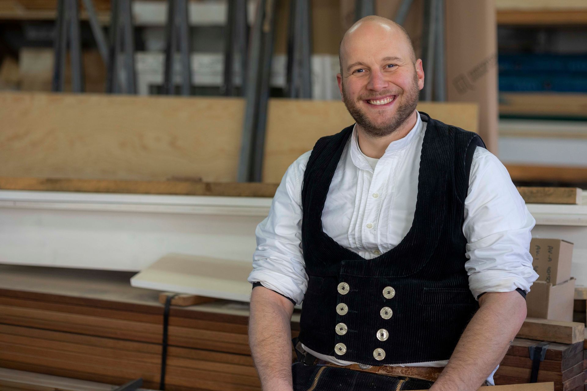 Ein lächelnder Mann in einer Werkstatt, bekleidet mit einer schwarzen Weste und einem weißen Hemd, ist von Holz umgeben.
