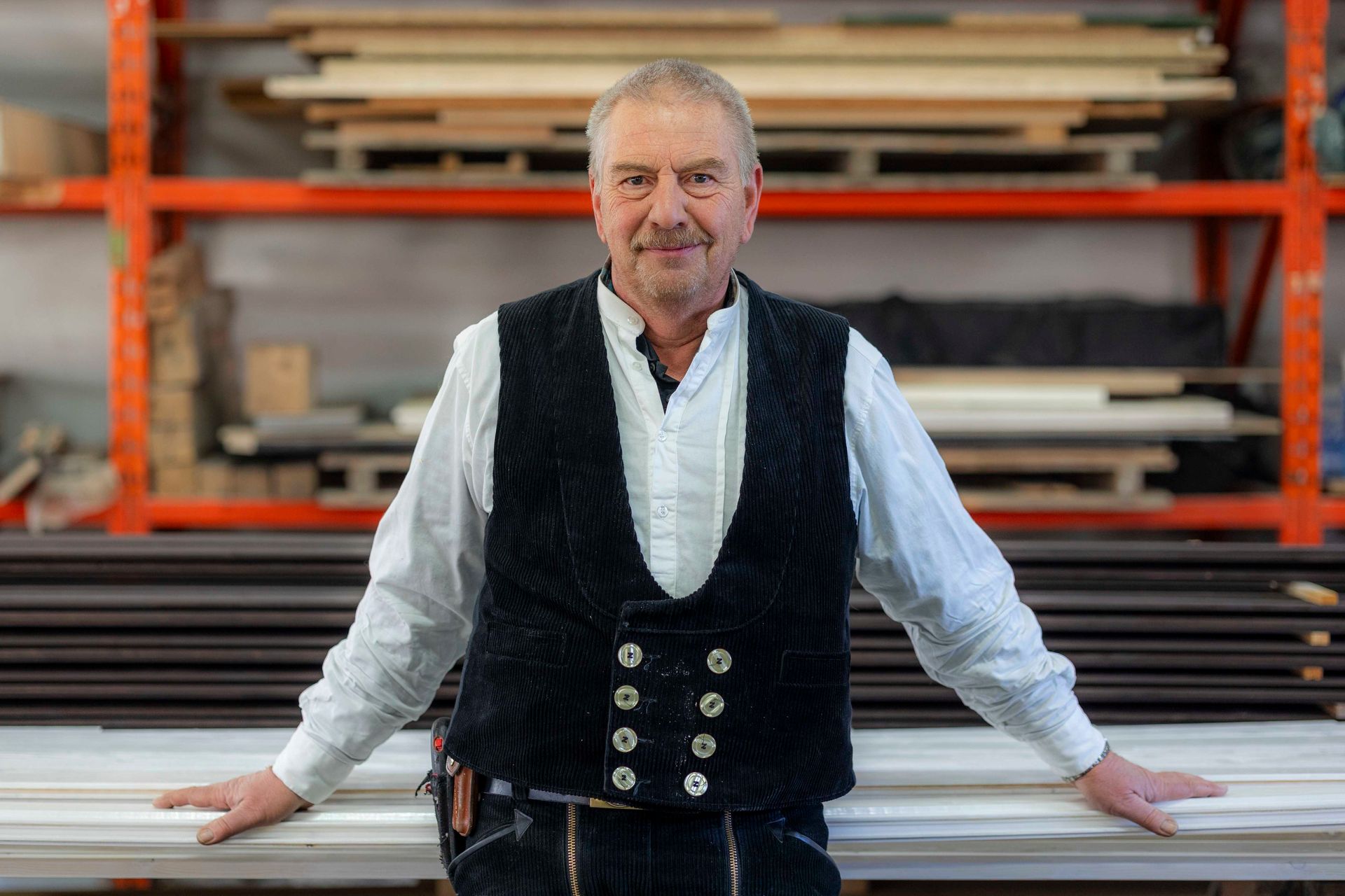 Mann in Arbeitsweste, lächelnd, die Arme auf Holzstücken abgestützt. Werkstattregale im Hintergrund.