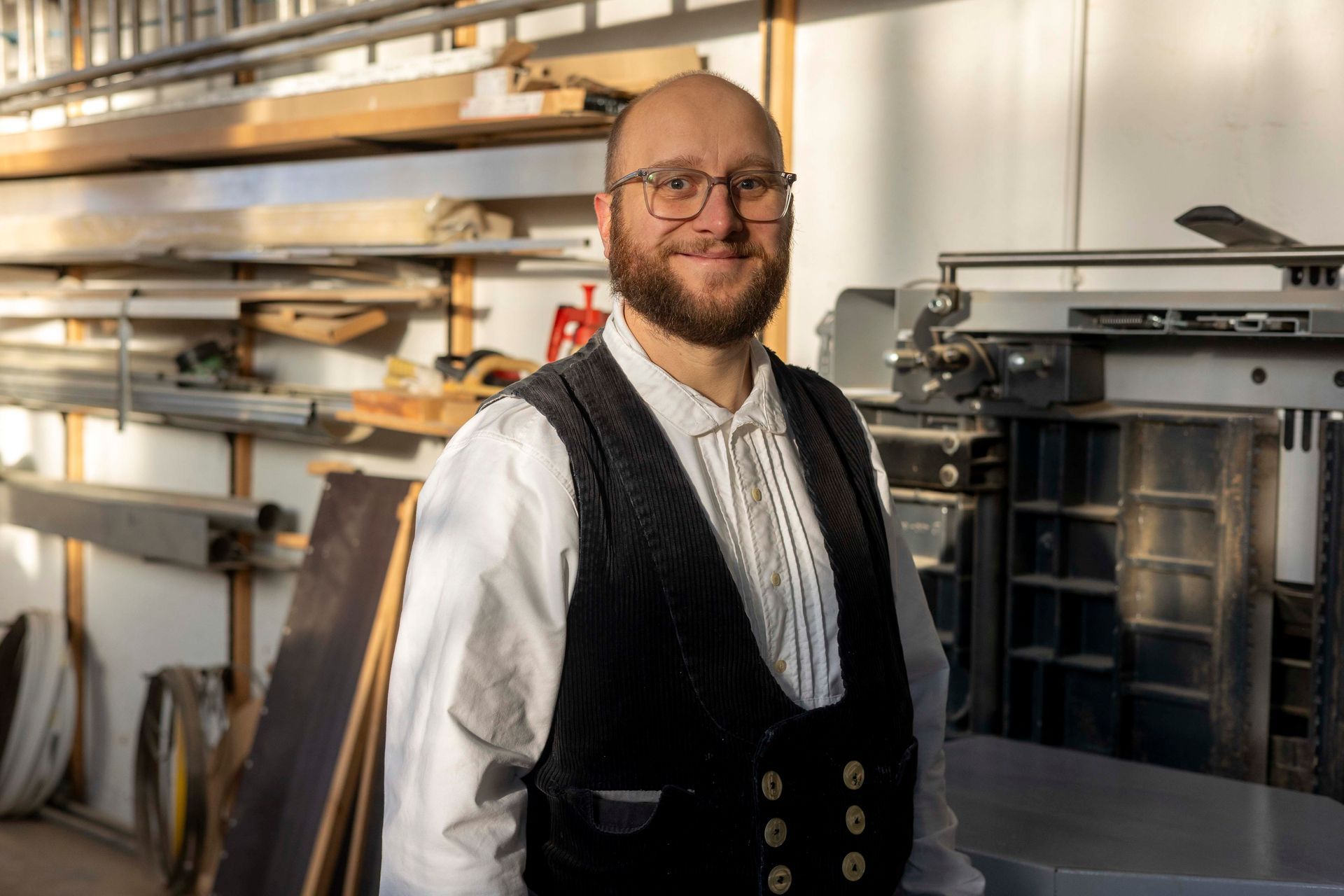 Bärtiger Mann in einer Werkstatt, mit Brille und schwarzer Weste, lächelt.