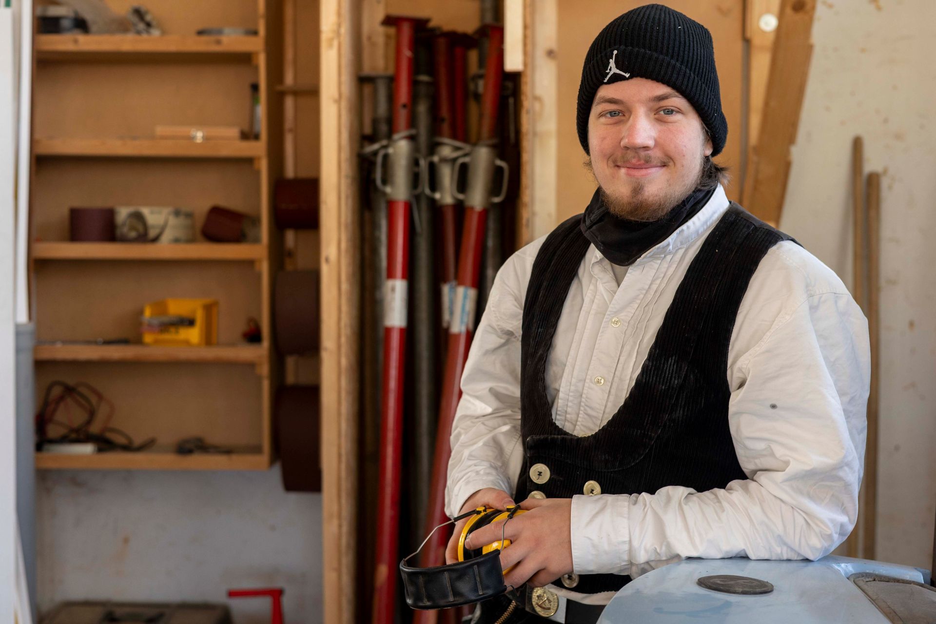 Mann in Arbeitskleidung mit Werkzeugen, lächelnd in einer Werkstatt mit Regalen und roten Stützen.