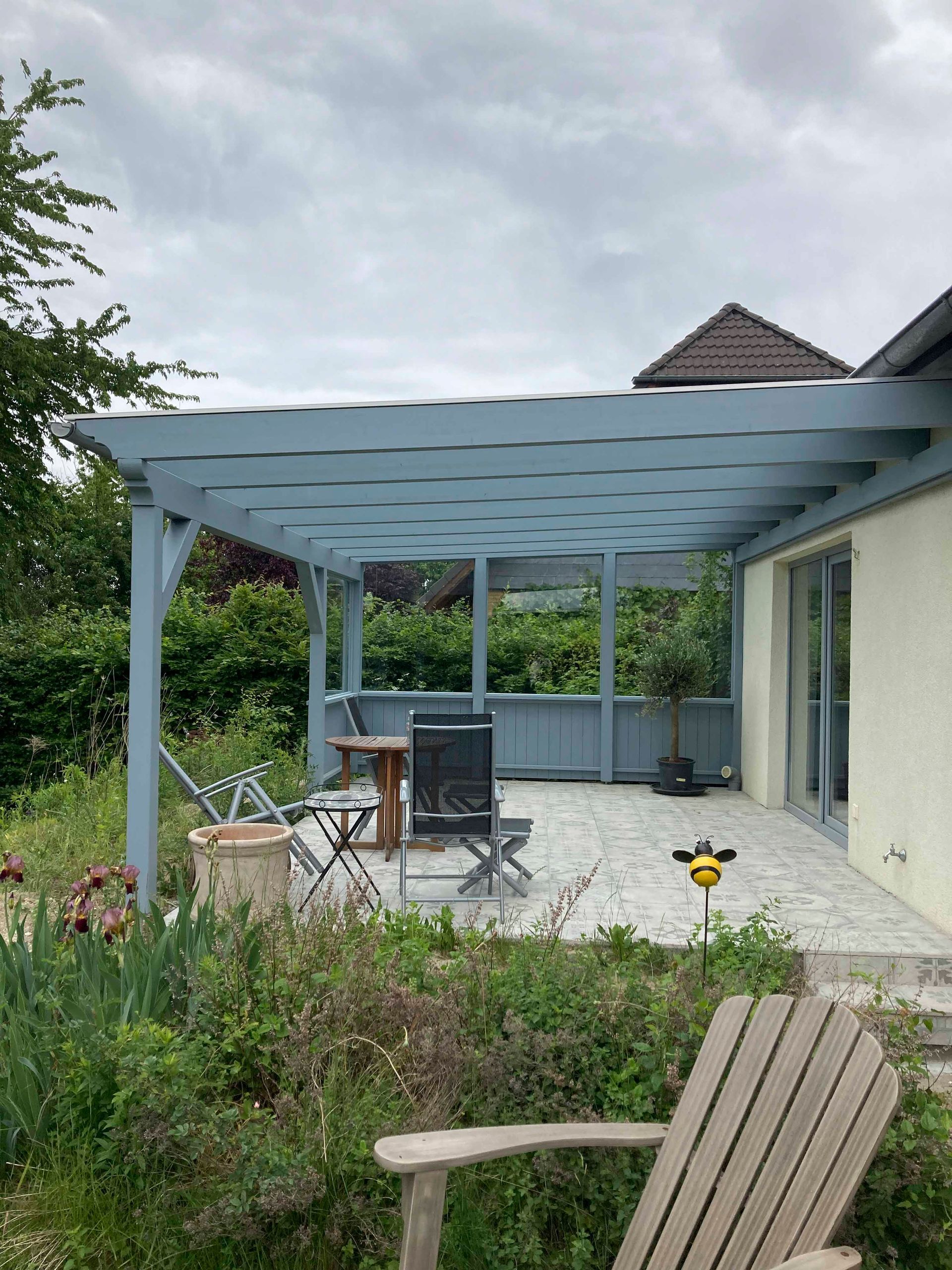 Eine blau gestrichene Pergola über einer Terrasse mit Gartenmöbeln, üppiges Grün im Vordergrund und ein Haus auf der rechten Seite.