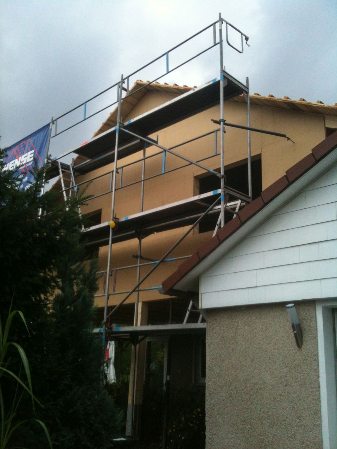 An einem zweistöckigen Haus mit beiger Fassadenverkleidung ist ein Gerüst errichtet, daneben ein weißes Haus mit rotem Dach. Bewölkter Himmel.