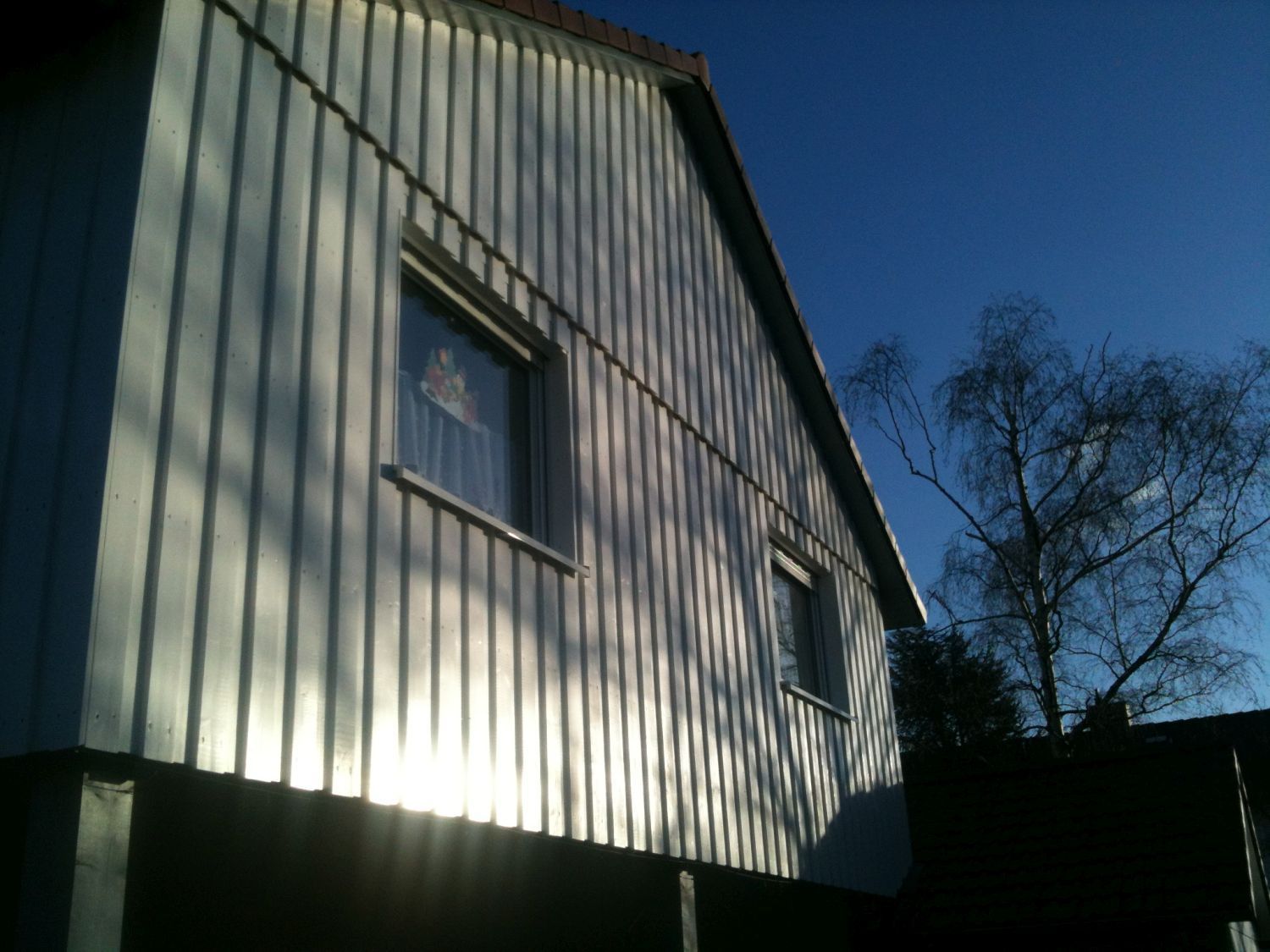 Außenansicht eines weiß getünchten Hauses mit zwei Fenstern, ein klarer blauer Himmel und ein kahler Baum.