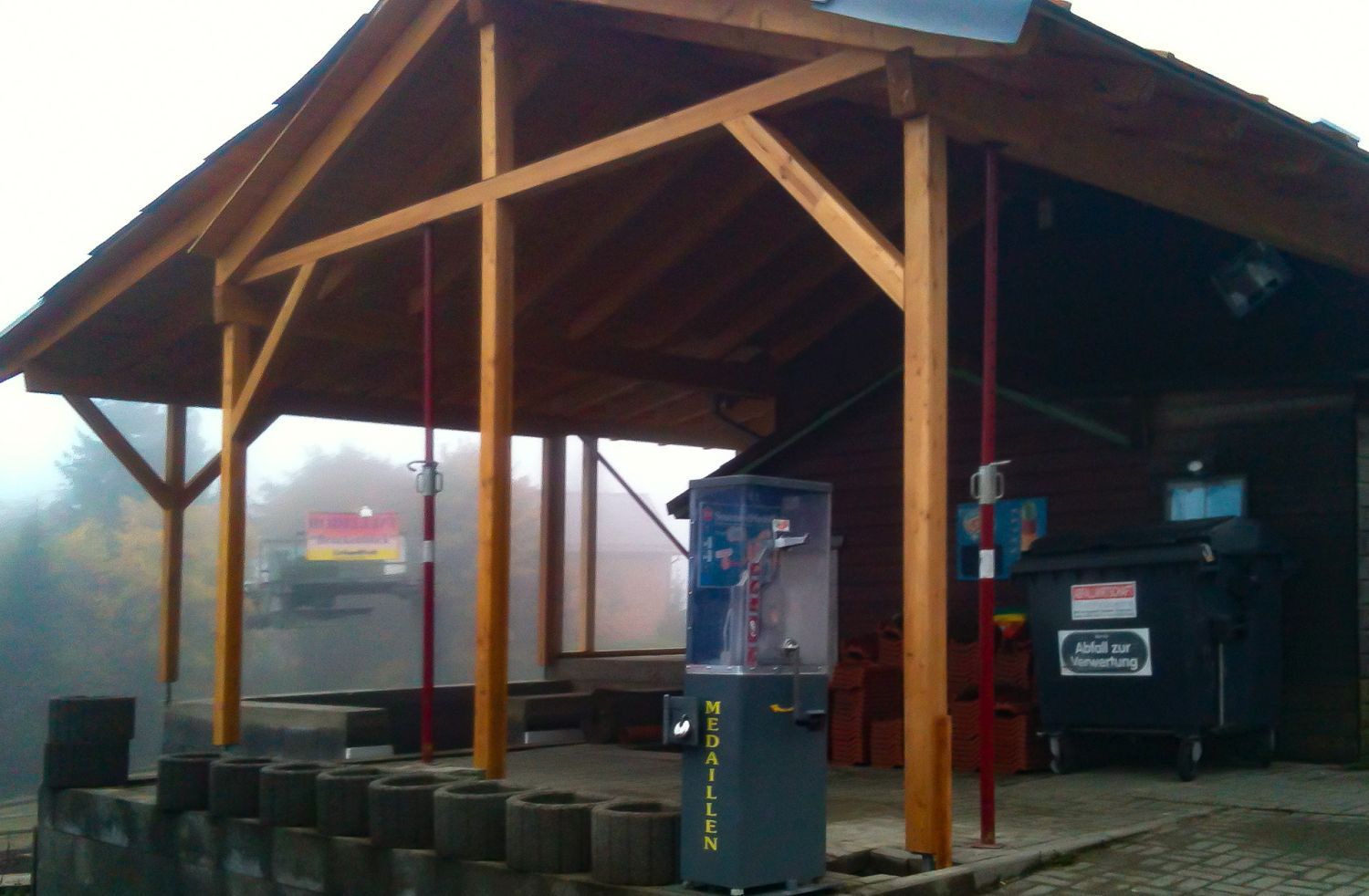 Holzkonstruktion mit roten Stützbalken, Mülleimer und Verkaufsautomat in nebliger Außenkulisse.