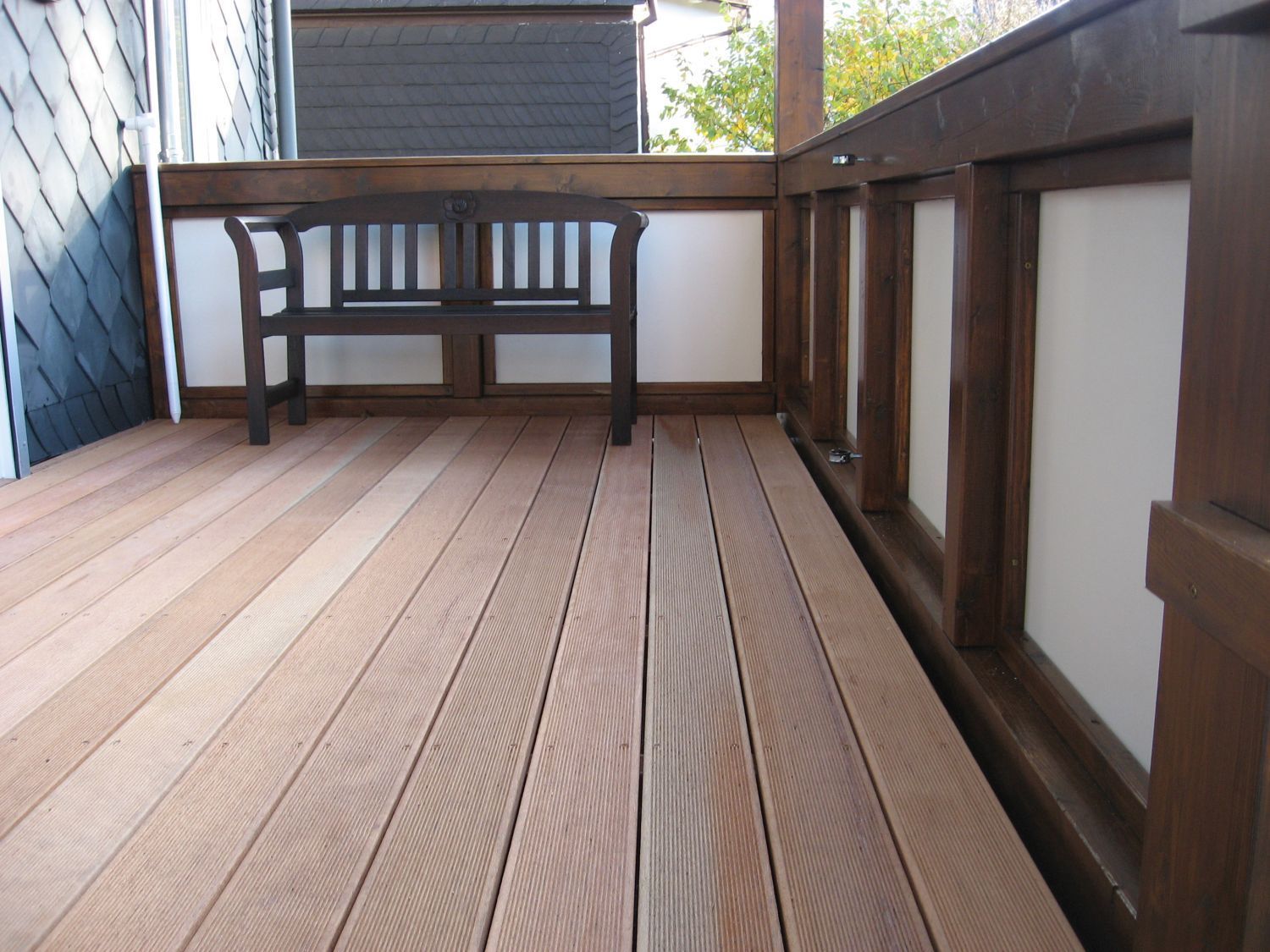 Holzterrasse mit brauner Bank, eingefasst von einem Geländer und weißen Paneelen.