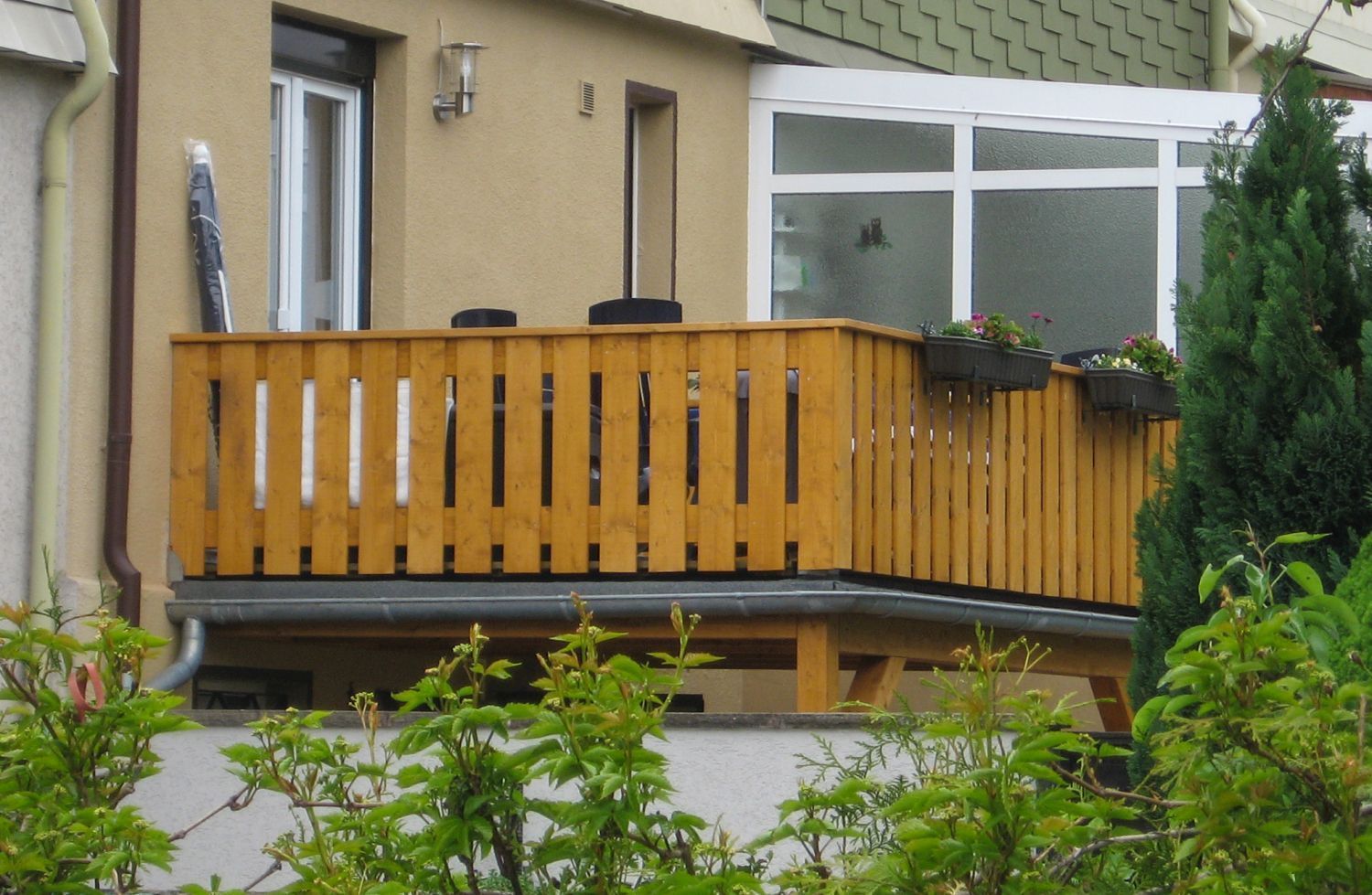 Ein hölzerner Balkon mit Geländer, der an ein helles Gebäude mit Fenstern angebaut ist. Im Vordergrund grüne Büsche.
