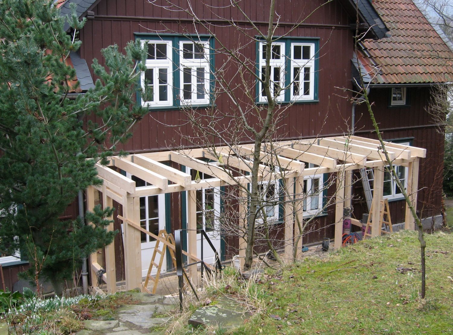 Holzrahmen eines im Bau befindlichen Wintergartens, der an ein braunes Haus mit weißgerahmten Fenstern angebaut ist.