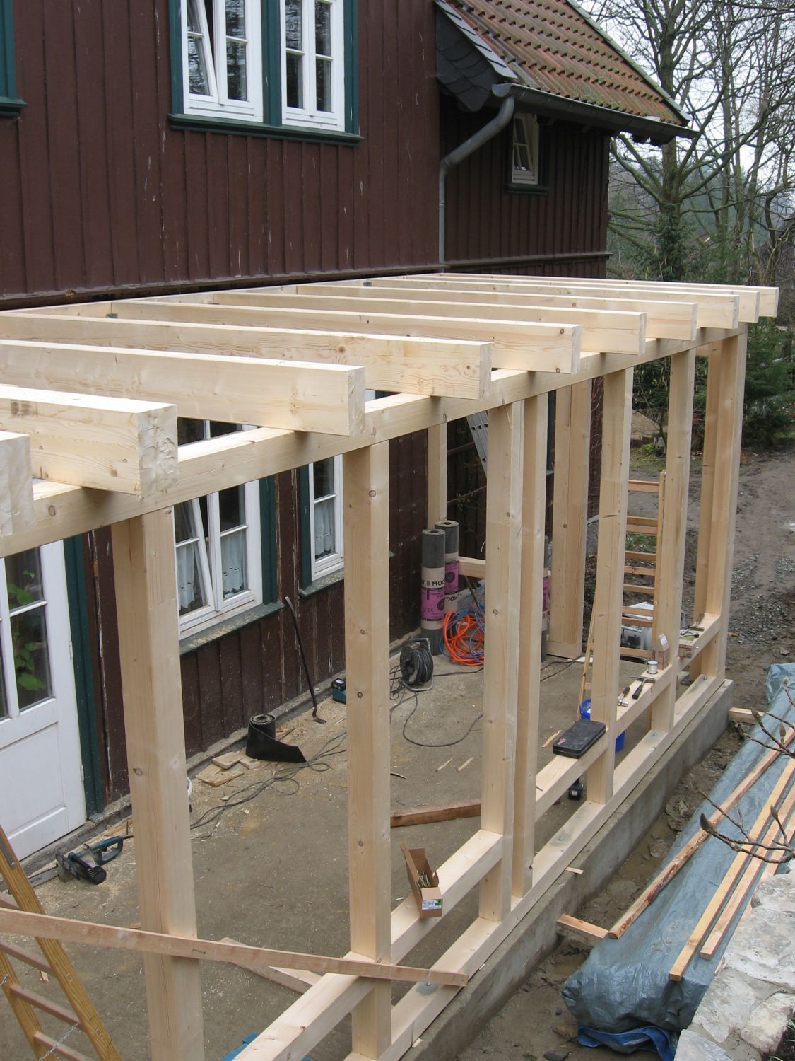 Holzgerüst einer im Bau befindlichen Veranda neben einem braunen Haus mit weißgerahmten Fenstern.