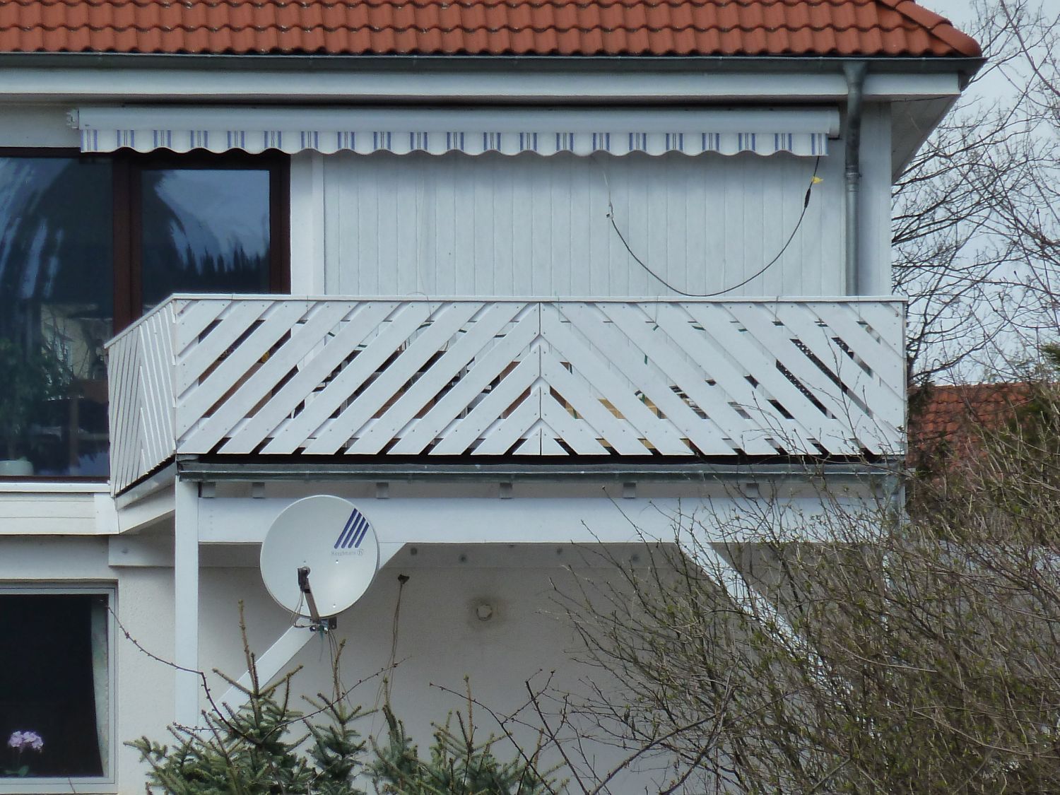 Weißer Balkon mit diagonalem Holzgeländer, Markise und Satellitenschüssel. Gebäude mit rotem Ziegeldach.