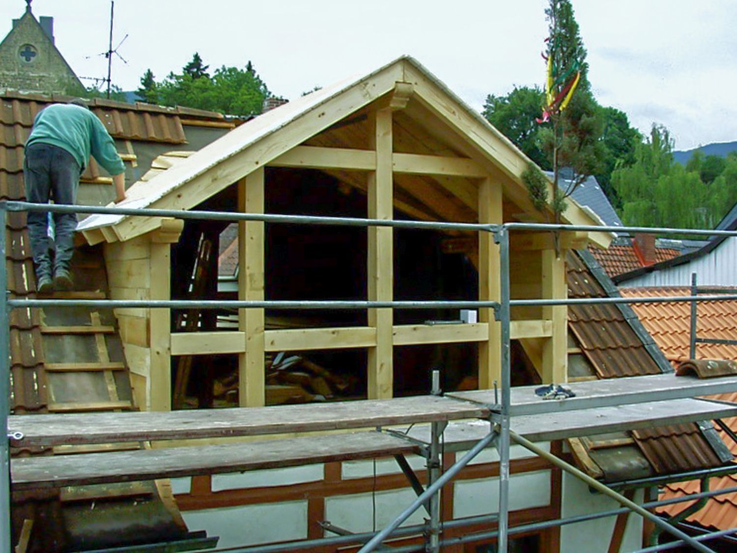 Ein Bauarbeiter montiert auf dem Dach die Holzkonstruktion eines Dachgaubenfensters. Gerüst, Dachziegel und Bäume sind zu sehen.