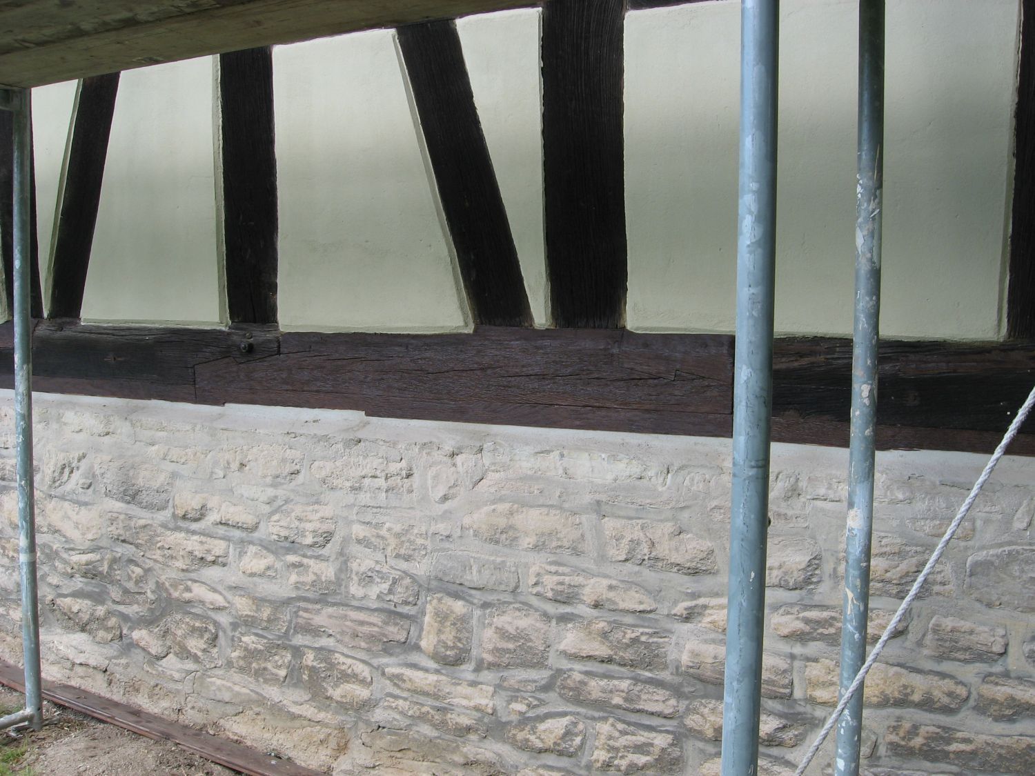 Steinmauer unterhalb dunkler Holzbalken und Milchglasscheiben, getragen von einem Metallgerüst.