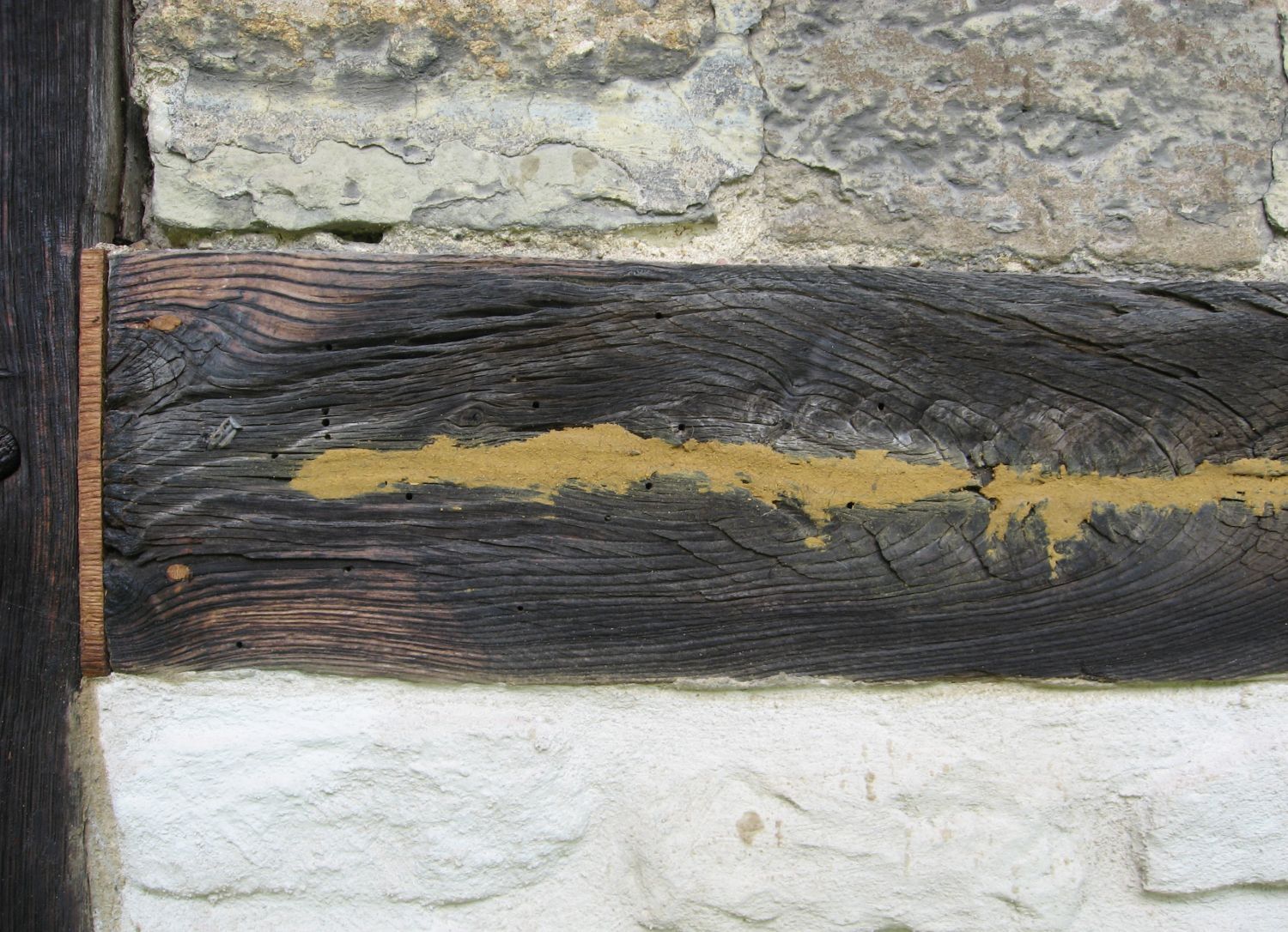 Dunkler Holzbalken mit gelber Füllung zwischen weißem und grauem Stein.