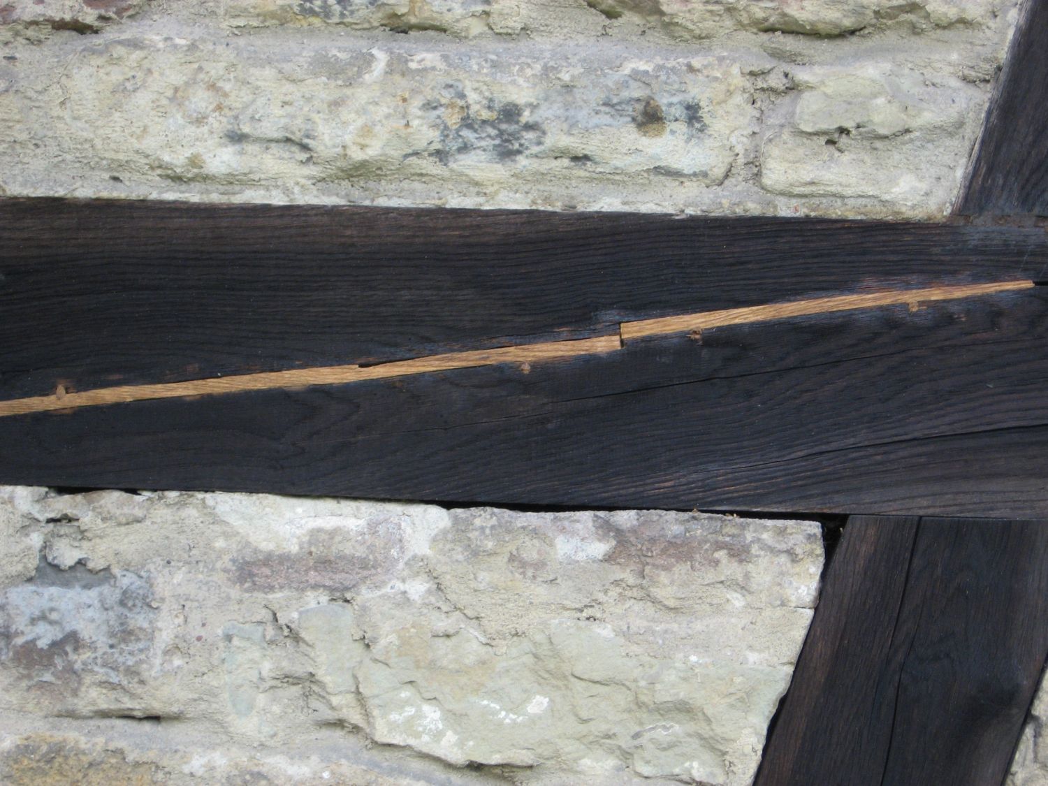 Dunkler Holzbalken in einer Steinmauer, mit einer sichtbaren, helleren Verbindungsstelle.