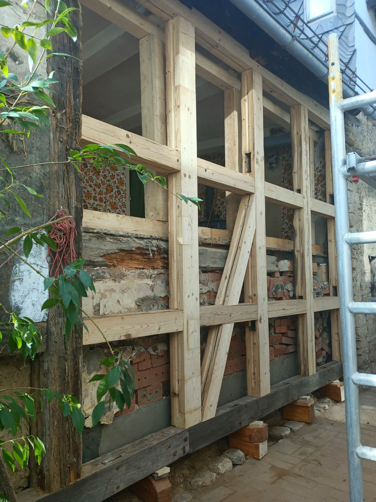 Holzrahmenkonstruktion über einer Ziegelmauer; eine Leiter steht daneben, Gebäudeaußenansicht.