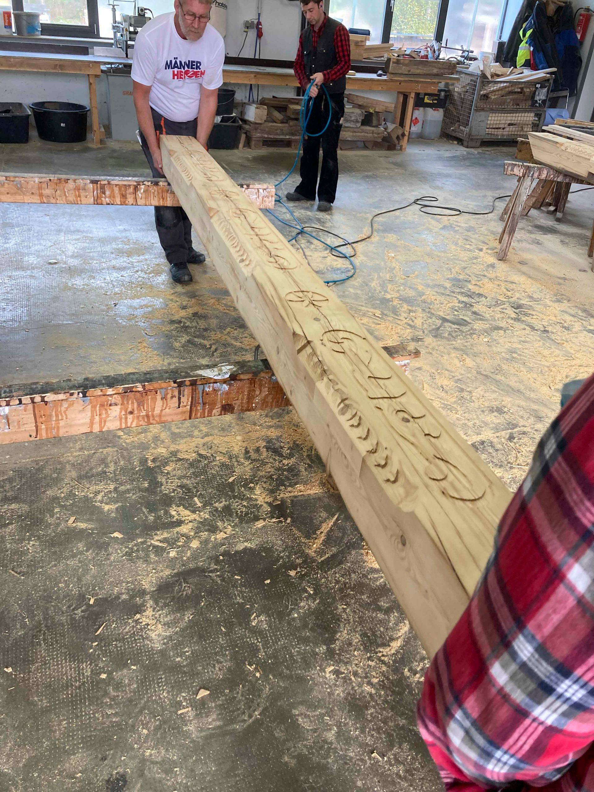 Ein Mann hebt einen großen Holzbalken an; Werkstattszene. Die Person benutzt eine Säge, um Linien in den Balken zu sägen.