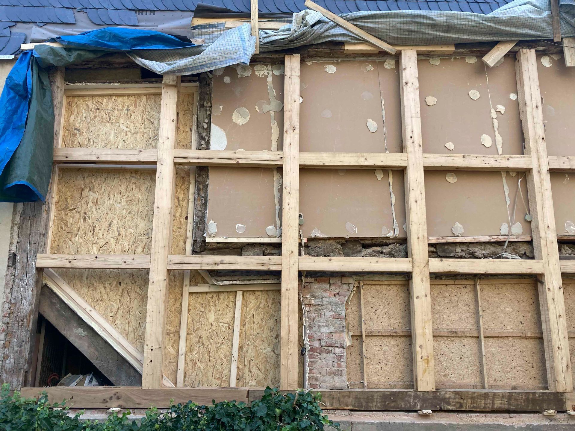 Außenwand im Bau, sichtbares Holzgerüst, verschiedene Paneele, blaue Plane darüber.