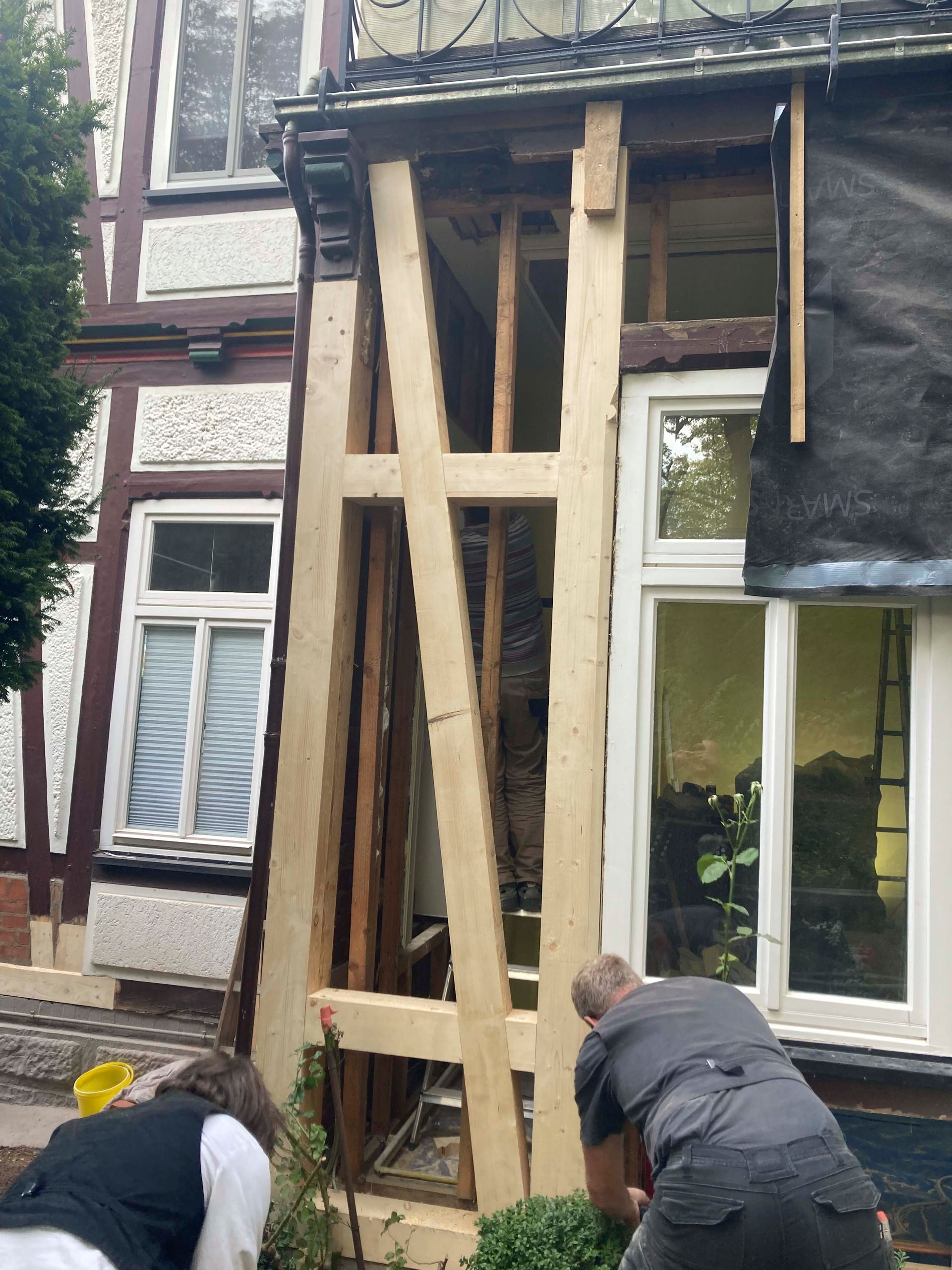 Außenansicht eines Gebäudes, auf dem ein Gerüst aufgebaut wird. Zwei Personen arbeiten, eine kniet.