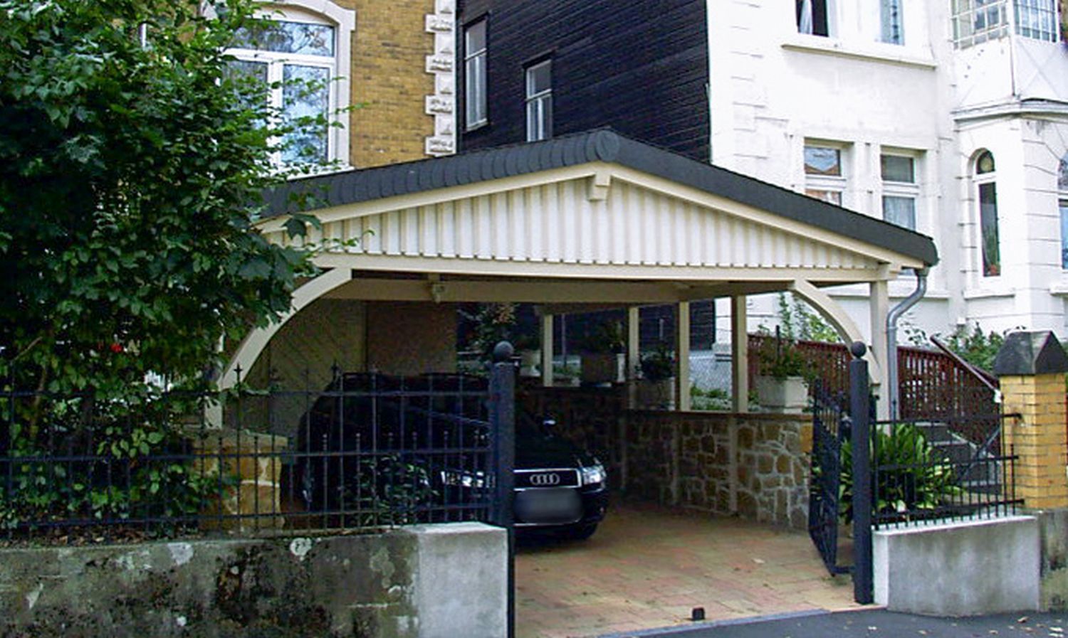 Ein an ein Gebäude angebauter Carport schützt ein schwarzes Auto. Er besteht aus einem Holzrahmen, einem Steinfundament und einem schwarzen Metalltor.