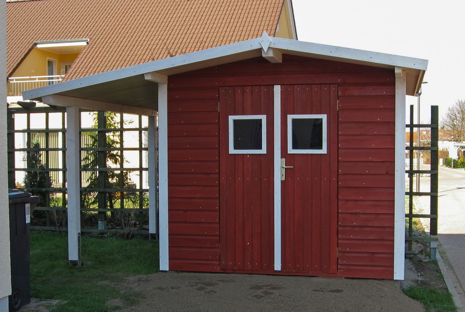 Roter Holzschuppen mit weißen Zierleisten und angebautem Carport.