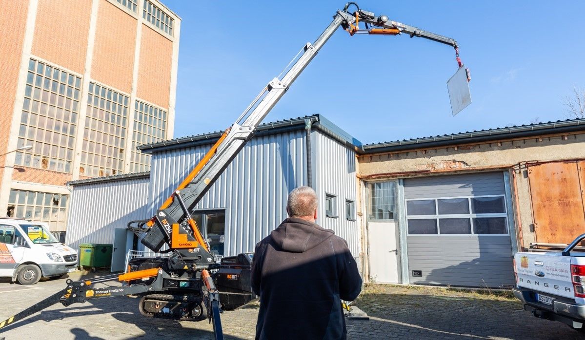 Ein Mann beobachtet, wie ein Mobilkran in der Nähe eines Lagerhauses eine große, reflektierende Platte anhebt.