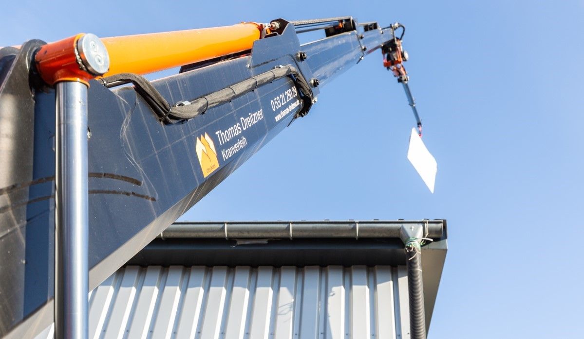 Ein Kranarm ragt über ein Gebäude und hält ein weißes rechteckiges Schild vor blauem Himmel.