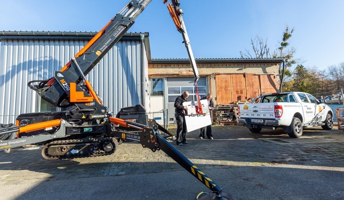 Ein Kran hebt ein Schild in der Nähe eines Gebäudes an; zwei Personen, ein Lastwagen und blauer Himmel sind zu sehen.