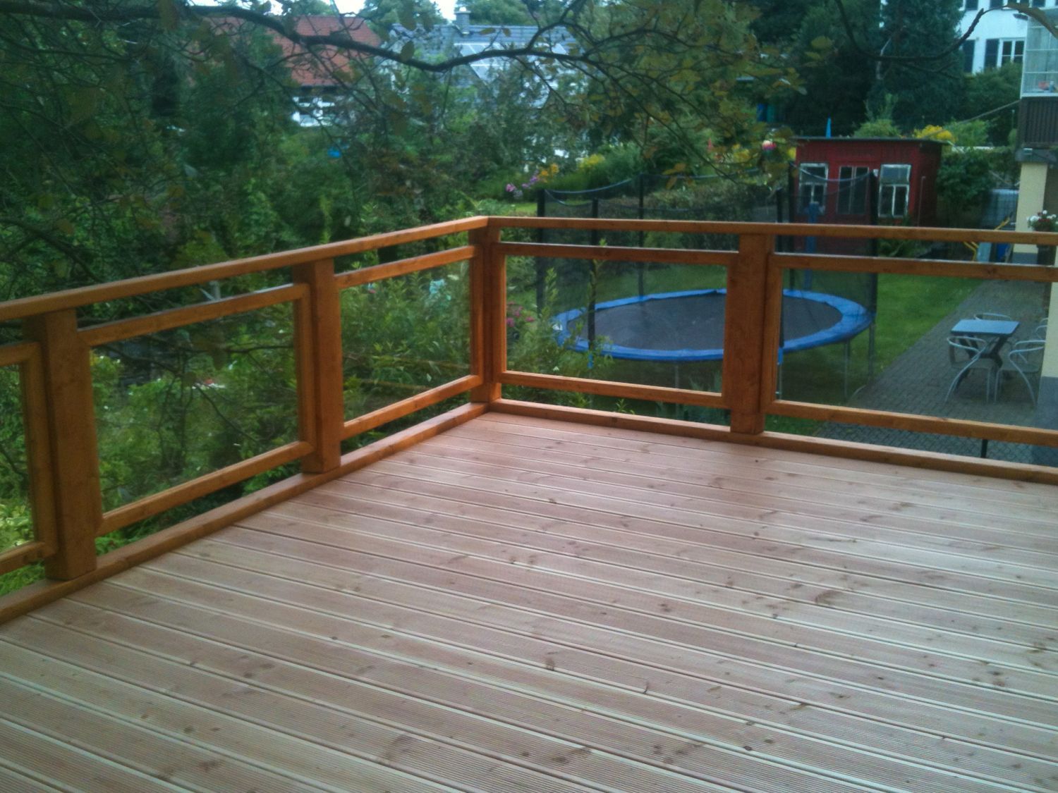 Holzterrasse mit Glasgeländer und Blick auf einen Garten mit Trampolin und Bäumen.