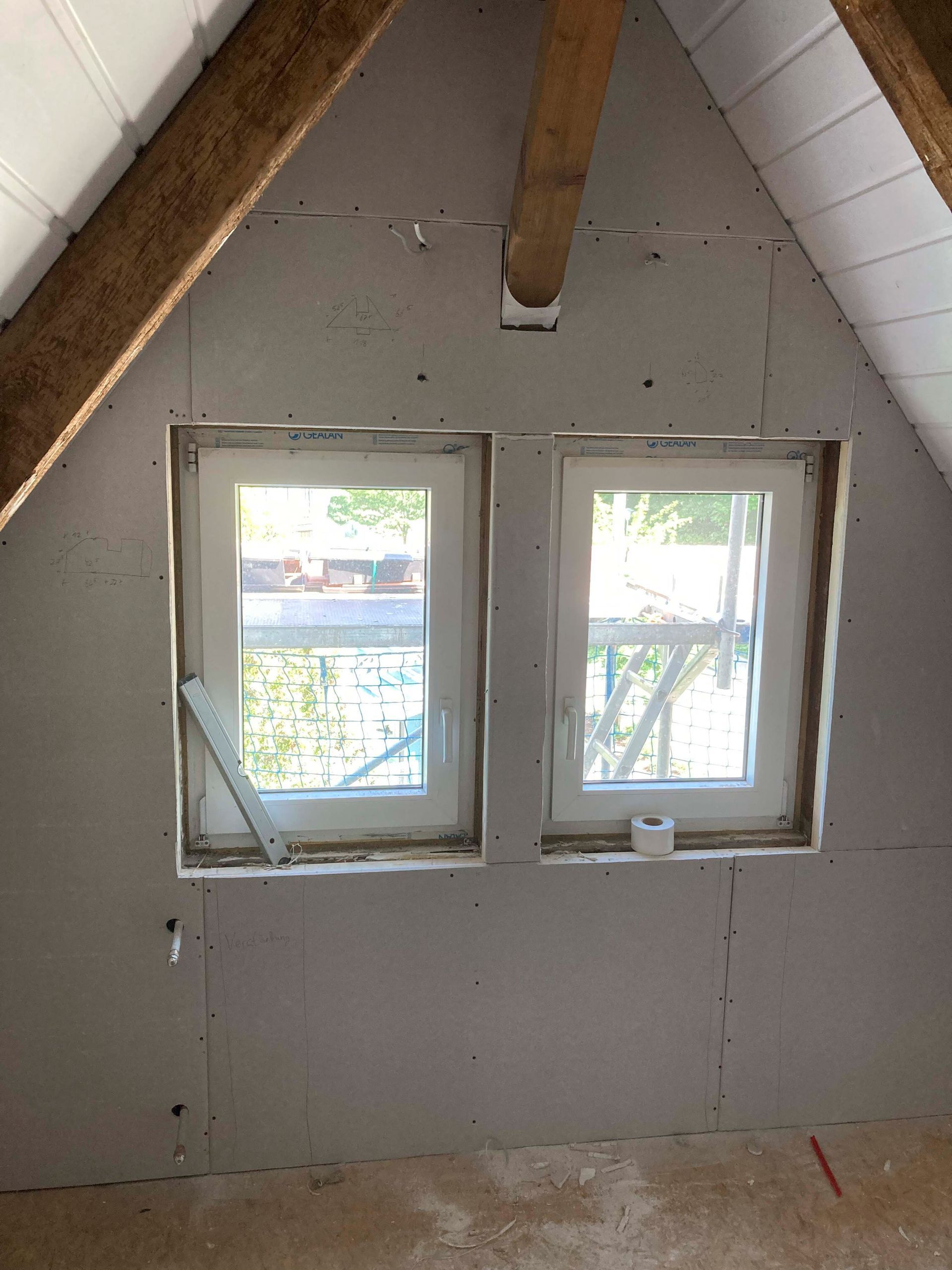 Innenansicht eines Zimmers mit zwei weißen Fenstern in einer Gipskartonwand. Darüber befinden sich Balken und eine Holzdecke.