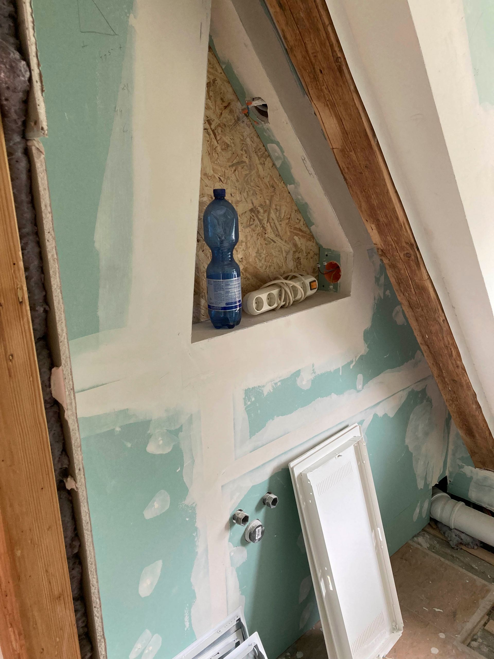 Ausbau im Dachgeschoss mit Gipskartonwänden und sichtbaren Holzbalken. Eine Wasserflasche steht in einer Nische.