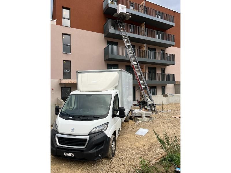 Une camionnette blanche avec une échelle attachée à l'arrière est garée devant un bâtiment.