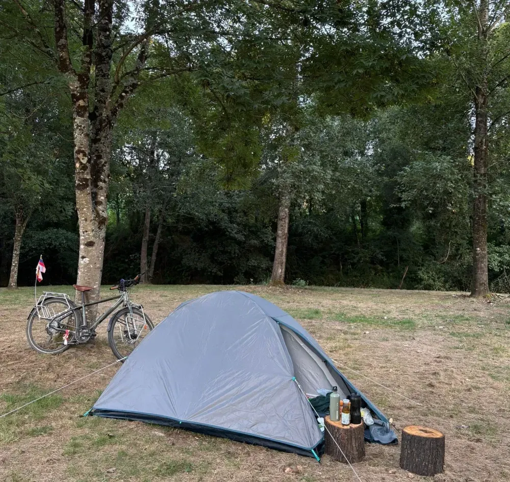 Tienda de campaña gris instalada en un camping arbolado junto a una bicicleta y equipo de acampada.