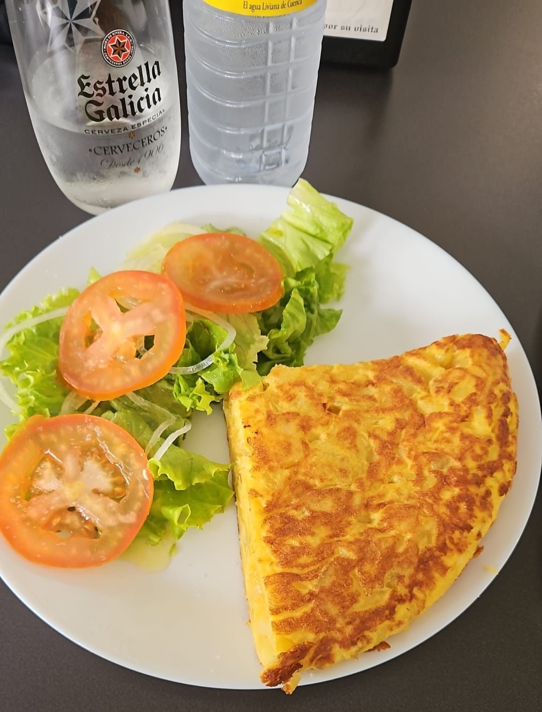 Un plato blanco con tortilla y tomates encima y una botella de agua Estrella Galicia.