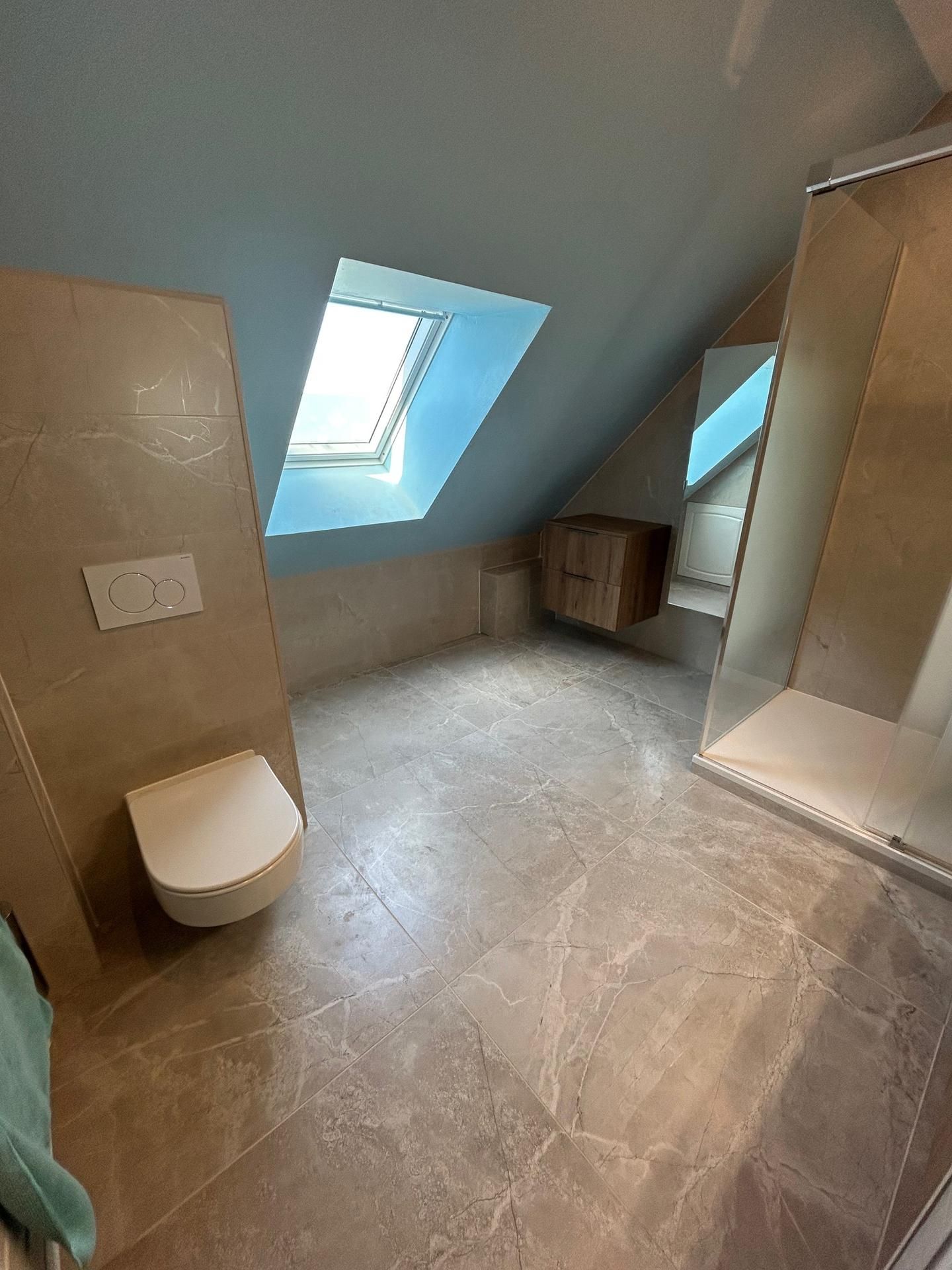 Salle de bains avec plafond mansardé, puits de lumière et carrelage en pierre. Toilettes suspendues, douche avec porte vitrée. Plafond bleu, tons neutres.