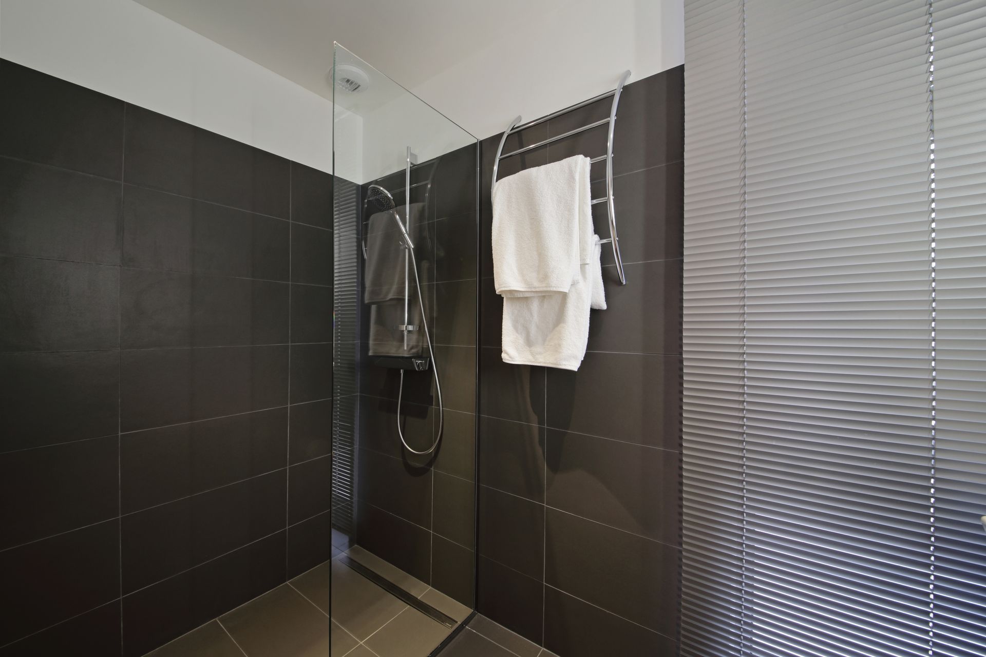 Douche carrelée foncée avec porte en verre et porte-serviettes contenant des serviettes blanches.