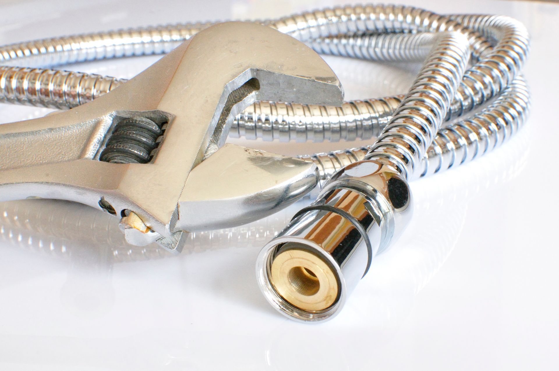 Clé tenant l'extrémité d'un tuyau flexible chromé, probablement destiné à la réparation de plomberie, sur fond blanc.